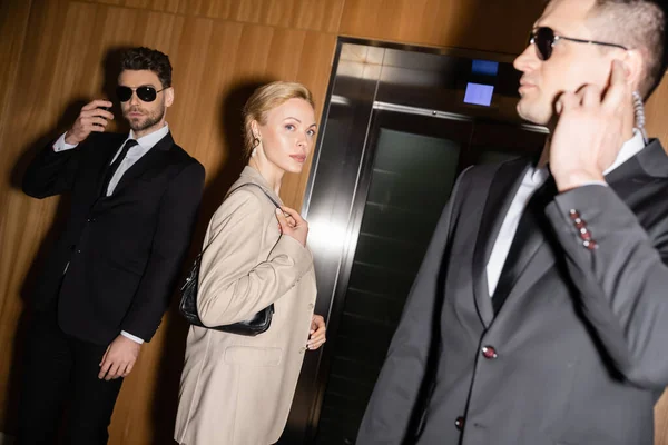 Personal security and protection concept, successful blonde woman with handbag standing near elevator next to bodyguards in suits and sunglasses, luxury hotel, female guest — Stock Photo