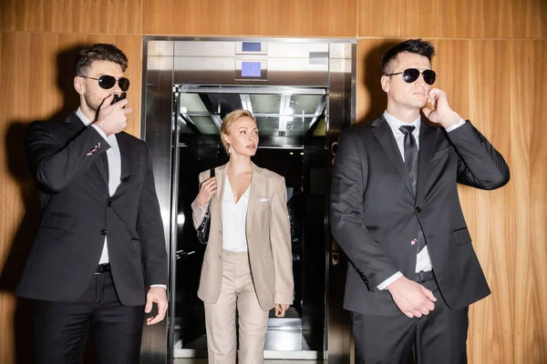 Persönliches Sicherheits- und Schutzkonzept, blonde und erfolgreiche Frau mit Handtasche, die aus dem Fahrstuhl steigt, Bodyguards in Anzügen und Sonnenbrillen, die ihre Privatsphäre im Luxushotel schützen — Stockfoto