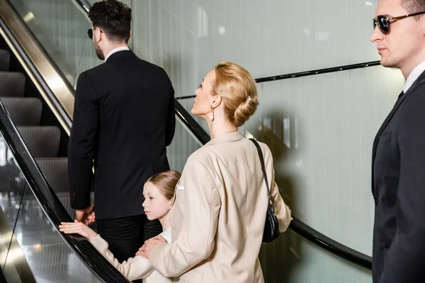 Personal safety, blonde and successful woman and preteen girl standing on escalator of luxurious hotel, two bodyguards protecting safety of clients, rich lifestyle, conceptual, professionals — Stock Photo