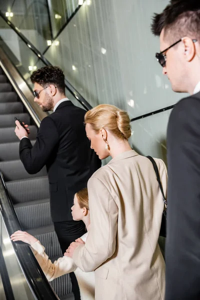 Private security concept, blonde and successful woman and preteen girl standing on escalator of luxurious hotel, two bodyguards communicating while protecting safety of clients, rich lifestyle — Stock Photo