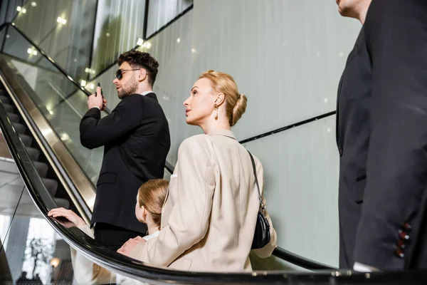 Personal security concept, blonde and rich woman and preteen daughter standing on escalator of hotel, two bodyguards communicating while protecting safety of clients, luxury lifestyle — Stock Photo