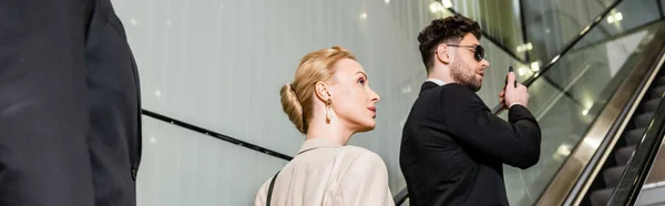 Personal security concept, professional bodyguards communicating while protecting safety of female client, rich lifestyle, blonde and successful woman standing on escalator of luxury hotel, banner — Stock Photo