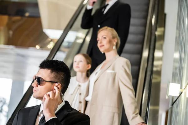 Personal security concept, bodyguard communicating through earpiece while protecting safety of clients, rich lifestyle, successful woman and preteen daughter standing on escalator of hotel — Stock Photo