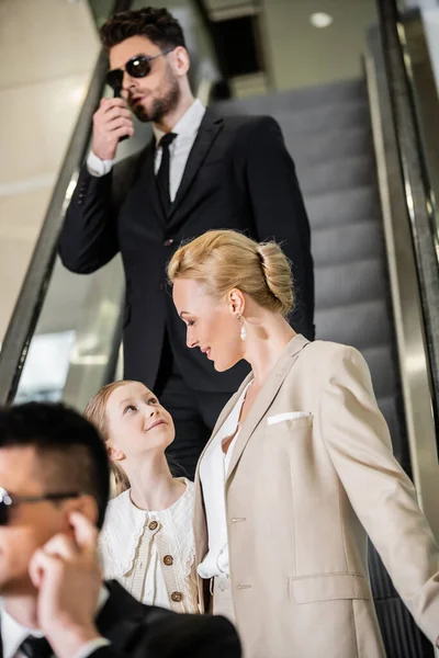 Concepto de seguridad personal, mujer rubia y feliz y preadolescente hija de pie sobre escaleras mecánicas de hotel de lujo, dos guardaespaldas que se comunican mientras protegen la seguridad de los clientes, rico estilo de vida - foto de stock