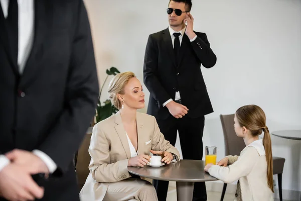 Private security concept, successful blonde woman and her preteen daughter spending time together in cafe, drinking coffee and orange juice, two bodyguards protecting safety of female clients — Stock Photo
