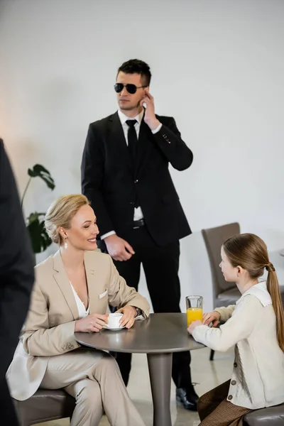 Private security concept, successful happy mother and daughter spending time together in cafe, drinking coffee and orange juice, two bodyguards protecting safety of female clients — Stock Photo