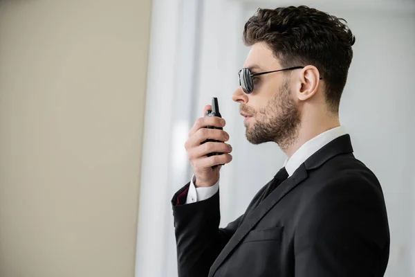 Surveillance, bodyguard communicating through walkie talkie, man in sunglasses and black suit with tie, hotel safety, security management, uniformed guard on duty, professional headshots, side view — Stock Photo