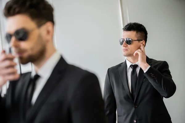 Bodyguard communicating through earpiece, man in sunglasses and black suit with tie, hotel safety, security management, surveillance and vigilance, working partner on blurred foreground — Stock Photo