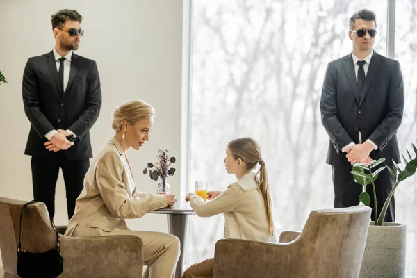 Private security concept, bodyguards standing near female clients, preteen girl and blonde mother spending time together in cafe, personal safety and protection, professional in suits — Stock Photo