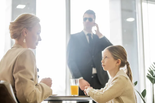 Preteen girl talking to blonde mother near bodyguard on blurred background, security service, private safety concept, glass of orange juice, personal safety — Stock Photo