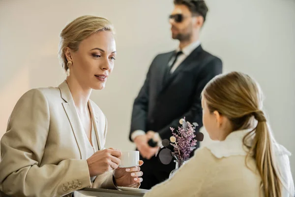 Blonde woman holding cup of coffee and talking to preteen daughter near bodyguard on blurred background, security service, private safety concept, quality time of mother and child — Stock Photo