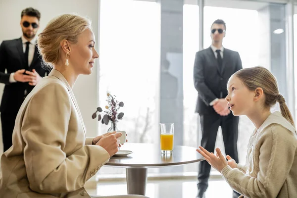 Security service, private safety concept, preteen girl talking with blonde mother over drinks in cafe, bodyguards standing on blurred background, personal safety, spending time together — Stock Photo