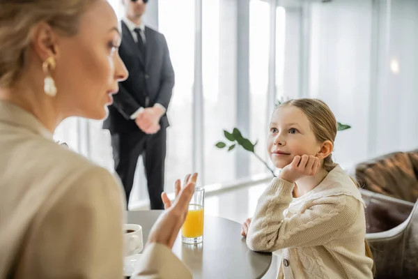 Preteen girl listening to blonde mother attentively, discussing something in cafe, bodyguard on blurred background, security service, private safety concept, glass of orange juice, personal safety — Stock Photo