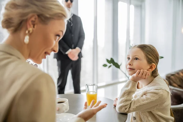 Preteen kid listening to blonde mother attentively, discussing something in cafe, bodyguard on blurred background, security service, private safety concept, glass of orange juice, personal safety — Stock Photo