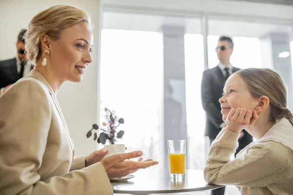 Security service, private safety concept, cheerful blonde woman with cup of coffee talking with preteen daughter in cafe, bodyguards on blurred background, beverages and communication — Stock Photo