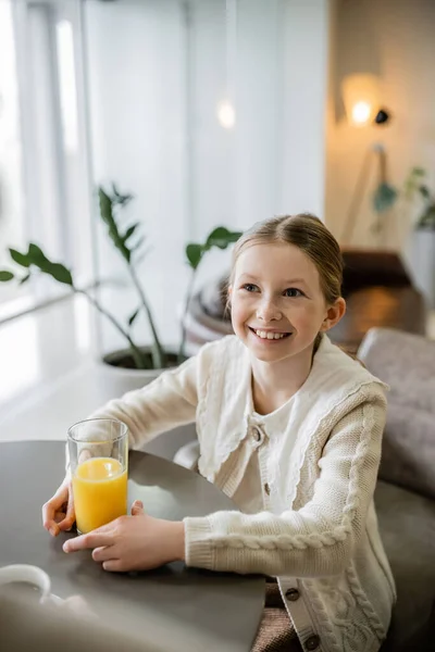 Happy preteen girl holding glass of orange juice while sitting in cafe, positivity and joy, round table, smart casual attire, blurred plant on background — Stock Photo