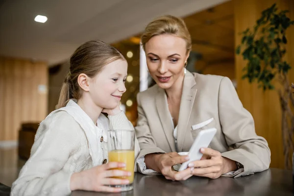 Счастливая мать и дочь проводят время вместе, блондинка держит смартфон рядом веселый дочь, цифровой возраст, работающий родитель и ребенок, современные родительские, семейные связи — стоковое фото