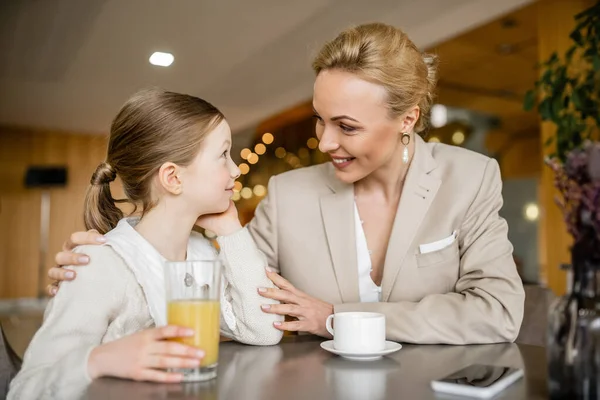 Happy mother spending quality time with daughter, blonde woman hugging preteen girl near beverages and smartphone, working parent and kid, modern parenting, family bonding — Stock Photo