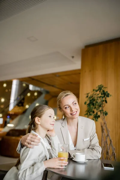 Happy mother and daughter spending quality time, blonde woman hugging preteen girl and looking away near beverages and smartphone, working parent and kid, modern parenting, family bonding — Stock Photo
