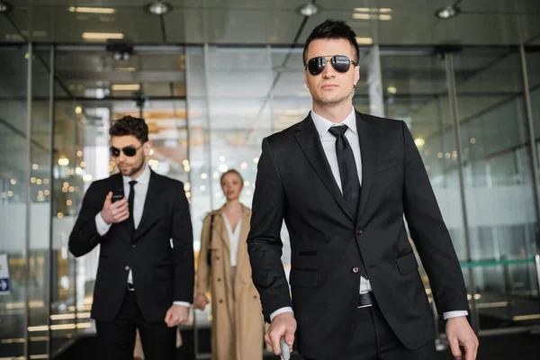 Private security service, personal safety concept, handsome bodyguards in formal wear and sunglasses protecting safety of client on blurred background, walking in hotel — Stock Photo