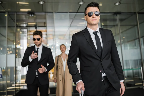 Private security service, personal safety concept, handsome bodyguards in formal wear and sunglasses protecting safety of blonde client on blurred background, men and woman walking in hotel — Stock Photo