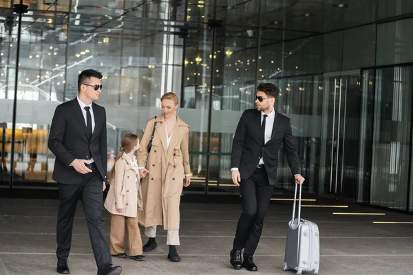 Bodyguards protecting privacy of female clients, woman and girl walking out of hotel on blurred background, personal safety of rich family, men in sunglasses and suit with luggage — Stock Photo
