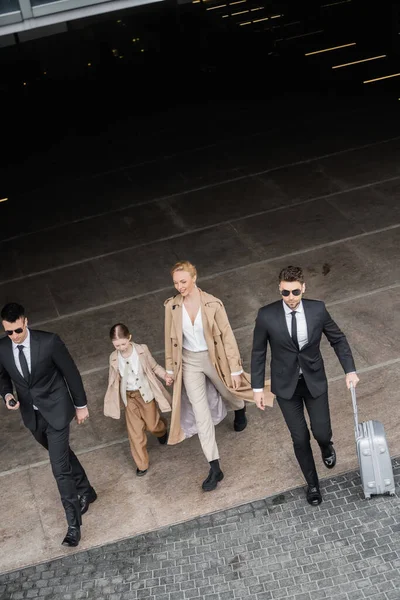 Private security, top view of personal bodyguards protecting female clients, blonde woman and daughter walking out of hotel, safety of rich family, men in formal wear and sunglasses on duty — Stock Photo