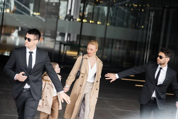 Handsome bodyguards protecting safety of female clients, blonde woman and daughter walking out of hotel, safety of rich family, men in sunglasses and suits on duty, work in security — Stock Photo
