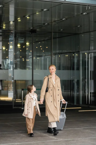 Autumn fashion, mother daughter time, happy woman with luggage holding hand of preteen girl while walking out of hotel together, smart casual, beige trench coats, outerwear, modern parenting — Stock Photo