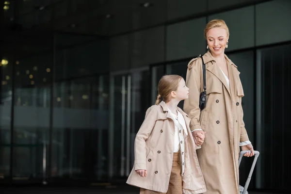 Autumn fashion, mother daughter time, positive woman with luggage holding hand of preteen girl while walking out of hotel together, smart casual, beige trench coats, outerwear, modern parenting — Stock Photo