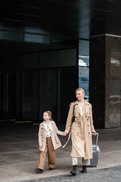 Moda de otoño, tiempo de la madre hija, mujer feliz con el equipaje cogido de la mano del niño preadolescente mientras que camina fuera del hotel juntos, casual elegante, abrigos de trinchera beige, ropa de abrigo, look de moda - foto de stock