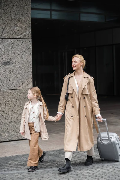 Outerwear, tempo da filha mãe, mulher feliz com bagagem segurando a mão da menina pré-adolescente enquanto saem do hotel juntos, casuais inteligentes, casacos de trincheira bege, moda de outono, parentalidade moderna — Fotografia de Stock