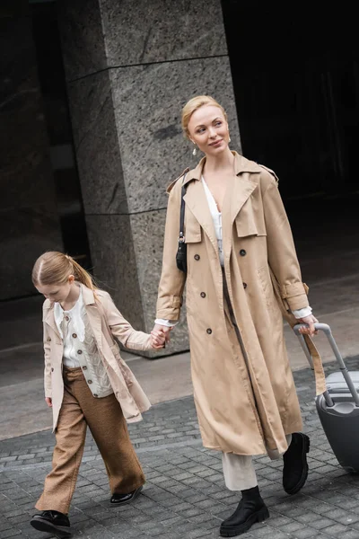 Modern parenting, mother daughter time, happy woman with luggage holding hand of preteen girl while walking out of hotel together, smart casual, beige trench coats, outerwear, trendy look — Stock Photo