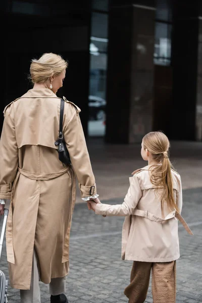 Modern parenting, mother daughter time, back view of blonde woman with luggage holding hand of girl while walking out of hotel together, smart casual, beige trench coats, outerwear, trendy look — Stock Photo