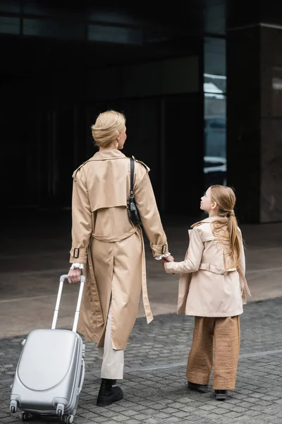 Family travel, mother daughter time, back view of blonde woman with luggage holding hand of girl while walking out of hotel together, smart casual, beige trench coats, outerwear, trendy look — Stock Photo