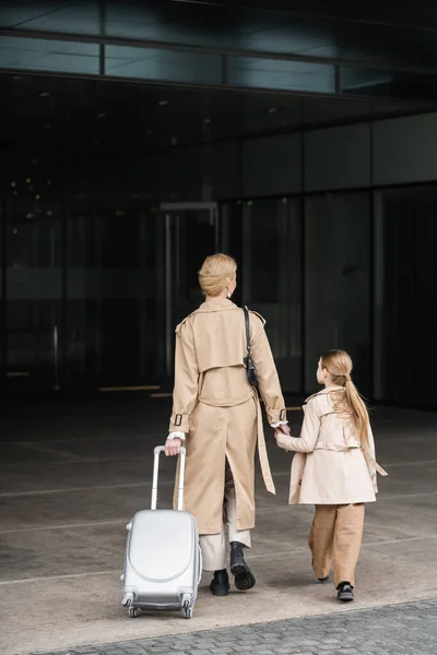 Family travel, mother and child, back view of blonde woman with luggage holding hand of girl while walking into hotel together, smart casual, beige trench coats, outerwear, trendy look — Stock Photo
