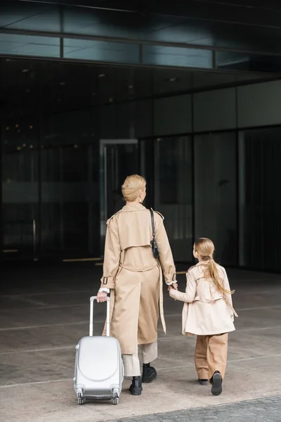 Family travel, mother and child, back view of blonde woman with luggage holding hand of girl while walking into hotel together, smart casual, beige trench coats, outerwear, smart casual — Stock Photo