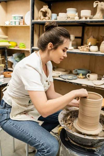 Vista lateral da artesã morena em avental trabalhando com argila na roda de cerâmica em estúdio em oficina — Fotografia de Stock