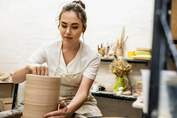 Brünette Handwerkerin in Schürze, die Tonvase herstellt und in der Keramikwerkstatt an der Töpferscheibe arbeitet — Stockfoto