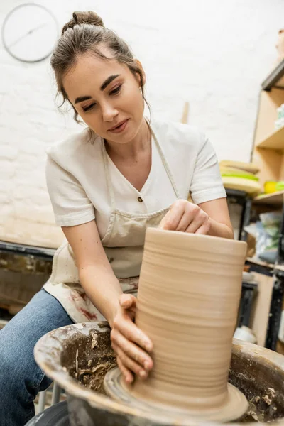 Morena alfarera femenina en delantal haciendo jarrón de arcilla en la rueda de cerámica en el taller de fondo - foto de stock