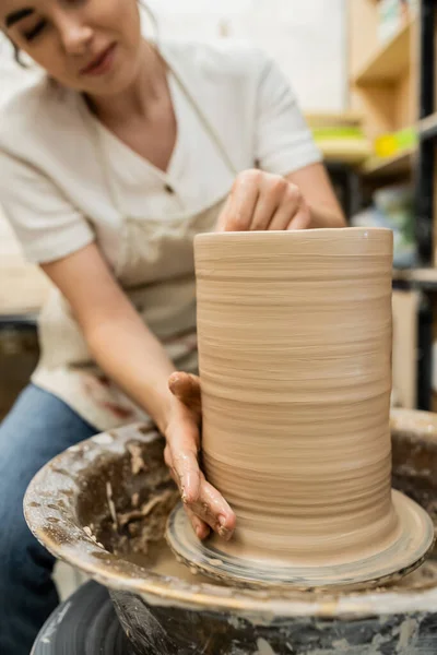 Vista recortada de alfarero hembra borrosa haciendo jarrón de arcilla en la rueda de cerámica en el taller en el fondo - foto de stock