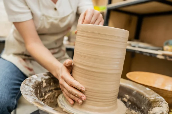 Обрізаний вид розмитого майстра в фартусі створення глиняної вази на гончарному колесі в майстерні — стокове фото