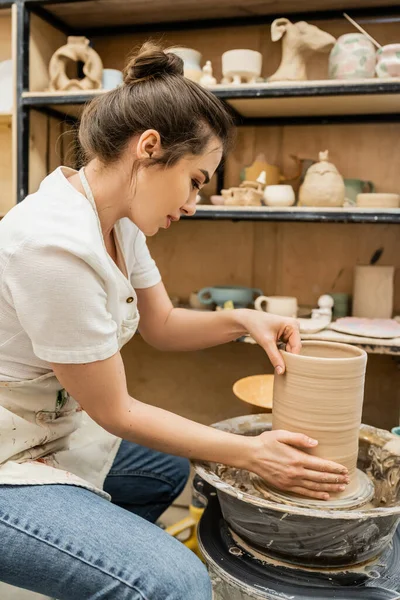 Vista lateral de la artesana en delantal trabajando con jarrón de arcilla sobre rueda de cerámica en estudio en taller - foto de stock