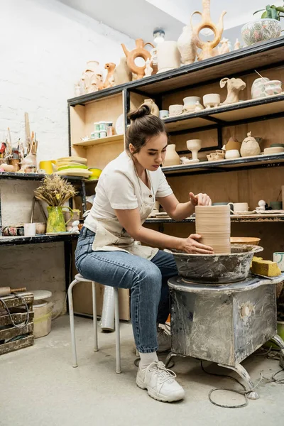 Брюнетка в фартуке создает глиняную вазу на гончарном круге в керамической мастерской — стоковое фото