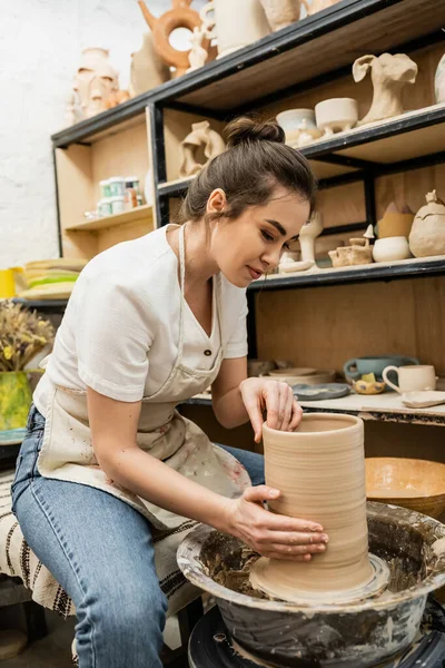 Брюнетка в фартуке создает глиняную скульптуру на гончарном круге в мастерской на заднем плане — стоковое фото