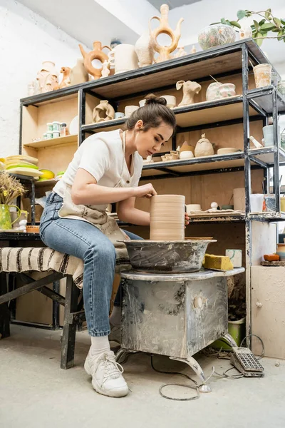 Brunette oleiro feminino em avental e vestuário de trabalho fazendo vaso de barro na roda de cerâmica no estúdio de cerâmica — Fotografia de Stock