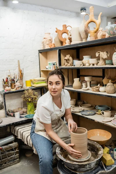 Brune artisanale dans un tablier regardant loin et faisant vase d'argile sur roue de poterie en atelier — Photo de stock