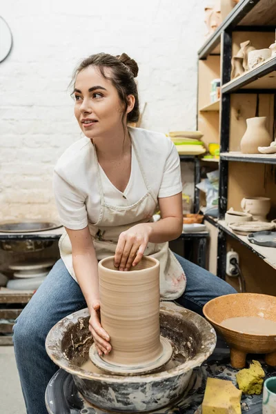 Sorrindo morena artesã em avental fazendo vaso de barro na roda de cerâmica na oficina — Fotografia de Stock