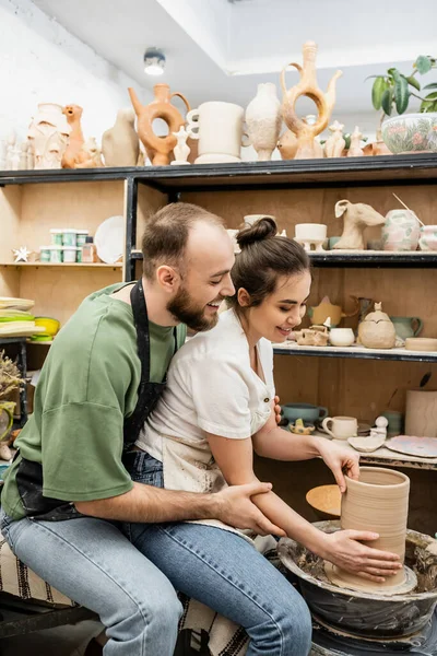 Smiling craftsman hugging girlfriend in apron while making clay vase on pottery wheel in workshop — Stock Photo