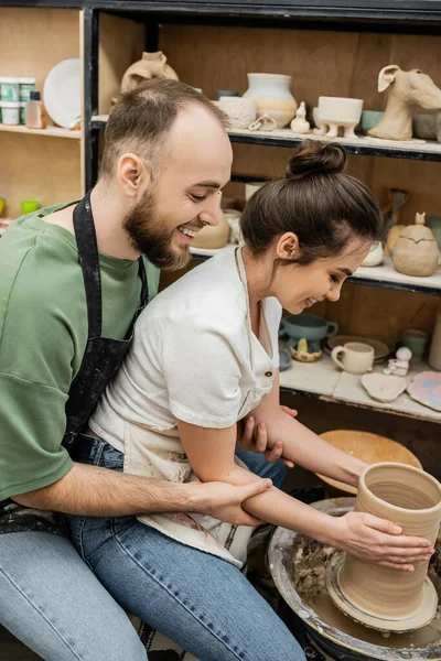 Side view of cheerful romantic artisans in aprons making clay vase on pottery wheel in workshop — Stock Photo